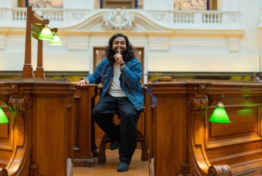 A person posing in the large wooden chair near the wooden partitions and green-shaded lamps inside the historic Domed reading room in State Library Victoria.