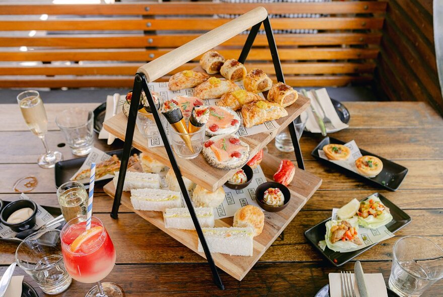 A two-tiered platter of savoury high tea snacks, plus cocktails and side plates, arranged on a wooden table. 