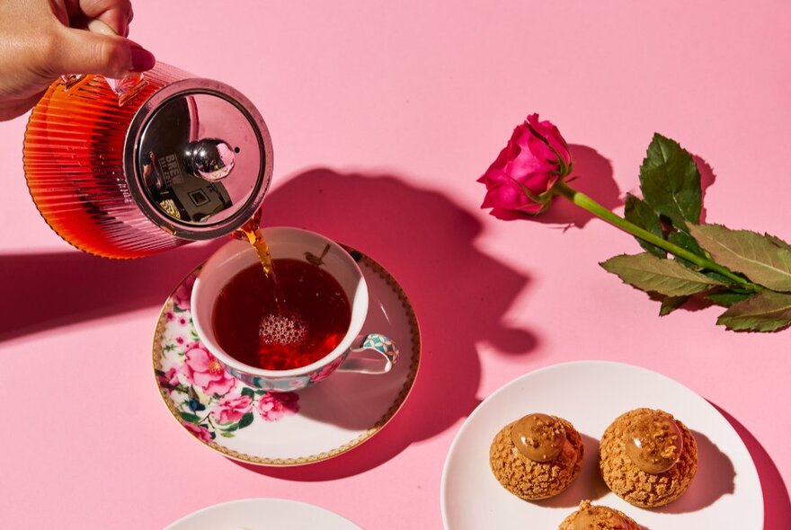 Teapot pouring tea into a china cup with biscuits on a saucer.