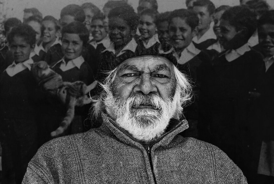 A black-and-white portrait of Uncle Kenny Ellwood Knight, an elderly Aboriginal man with a full white beard and a serious expression, positioned in front of a historical photograph of a group of smiling children in school uniforms.