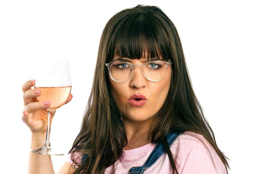 A female comedian wearing glasses, a pink t-shirt and holding up a glass of rose-coloured wine.