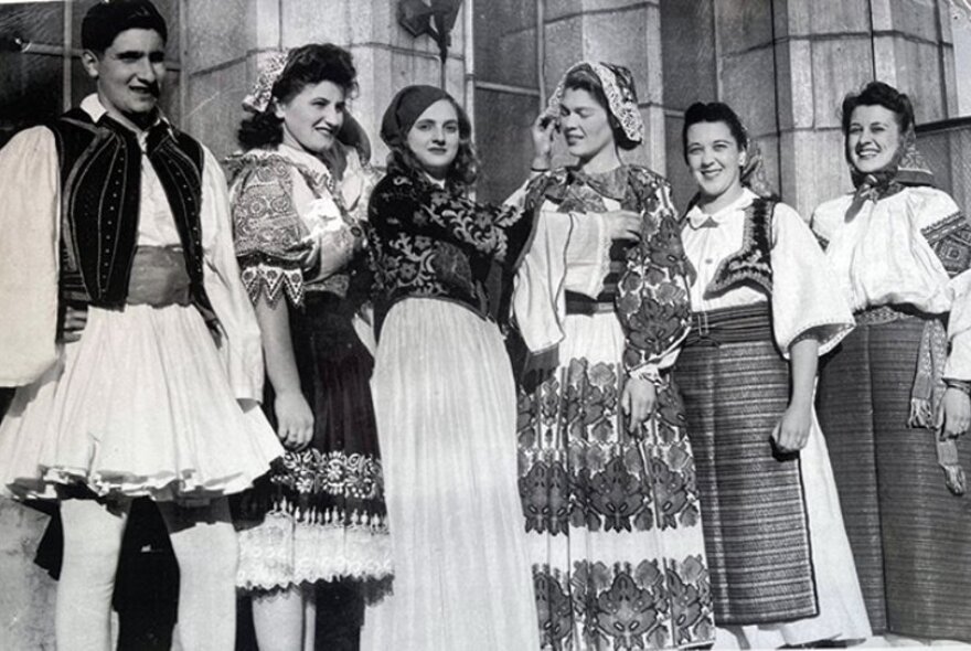A black and white photograph of five beautiful Greek women and one man in traditional Greek dress, lined up in front of a stone building.