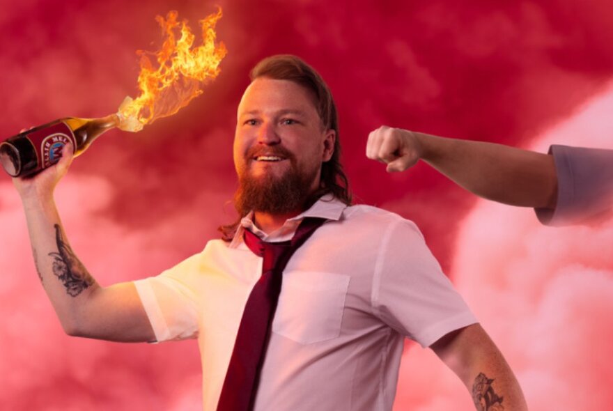Comedian Brett Black smiling while holding up a lit molotov cocktail with a hand reaching out to punch him in the head from behind.