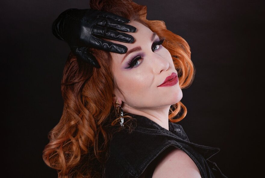 A woman with red hair, looking back at the camera over her shoulder with one gloved hand over her forehead.