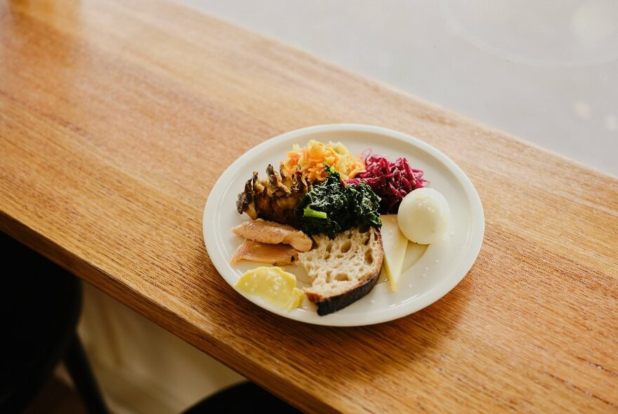 Plate of food that includes a slice of bread, pickles, cheese and dips on a timber counter.