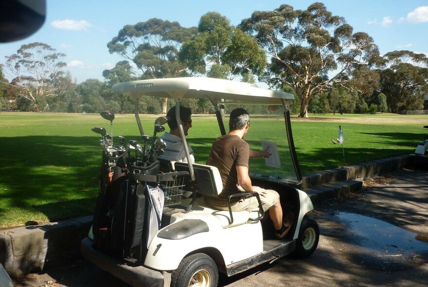 Two people driving past a golf course green in a golf cart, with two golf bags on the back.