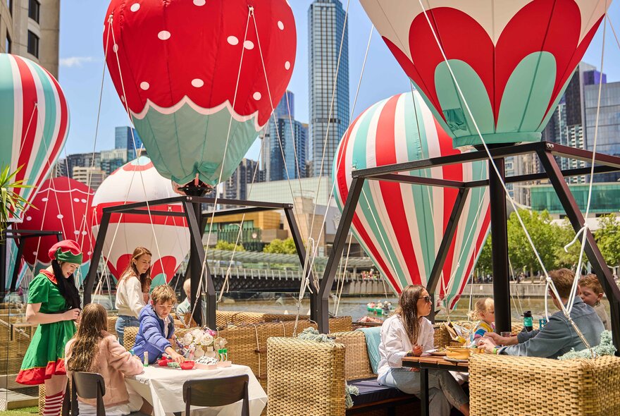 Outdoor dining spaces set up with basket booths and mini hot air balloons suspended above the tables, with people seated and enjoying food and drinks. The Yarra River and city buildings in the background.