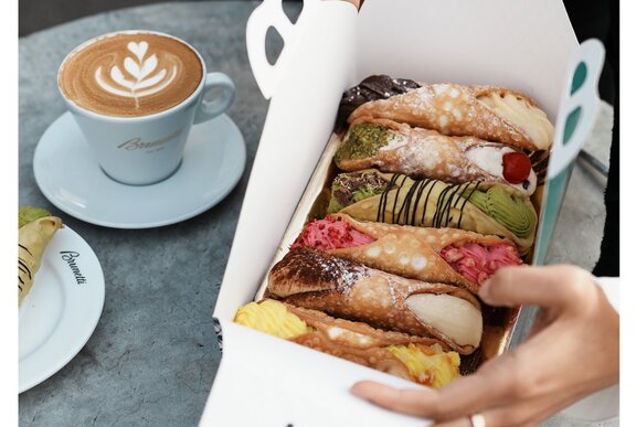 Hands opening a cannoli tasting box with a flat white coffee in a white cup and saucer