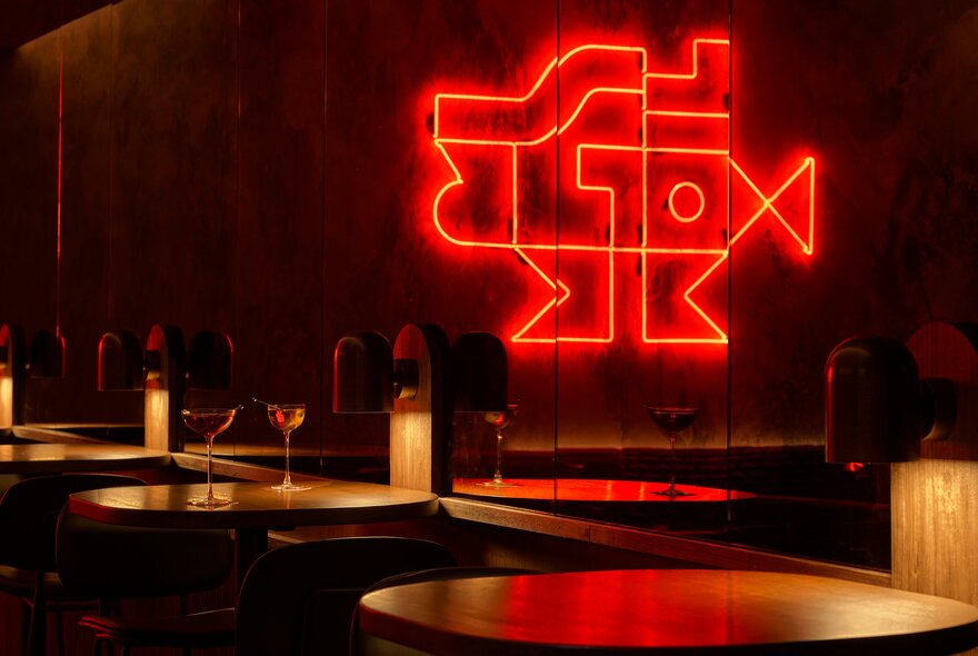 Warmly lit interior of Mr Mills bar, with small  tables arranged against a wall and the reflection of a red neon artwork on the wall.