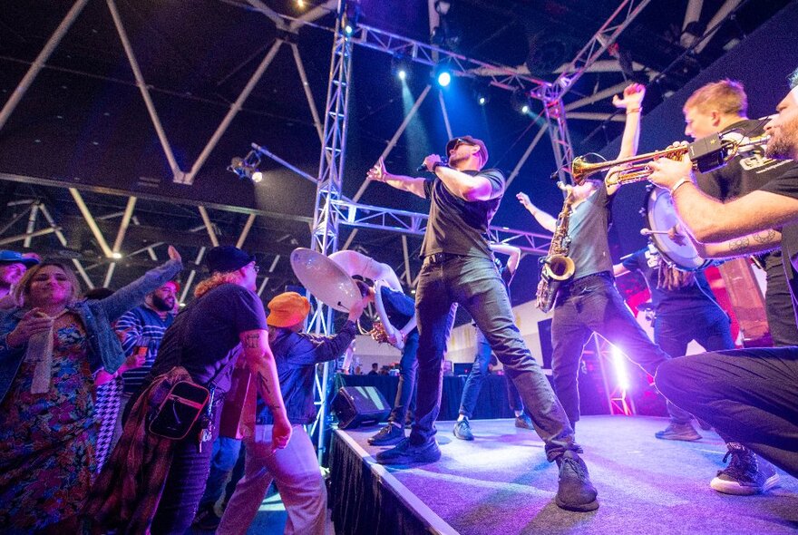 Brass band with singer performing on a purple-lit stage in front of a small crowd.