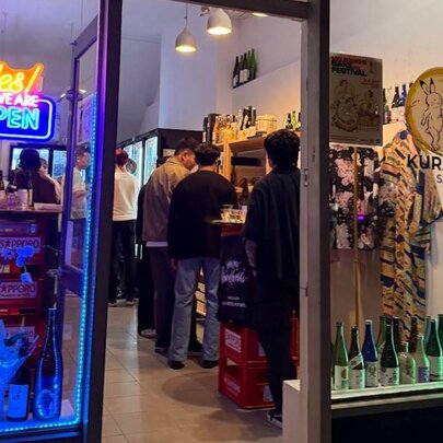Small Japanese liquor shopfront and bar with neon signs, bottle displays and people seated at the bar and tables.