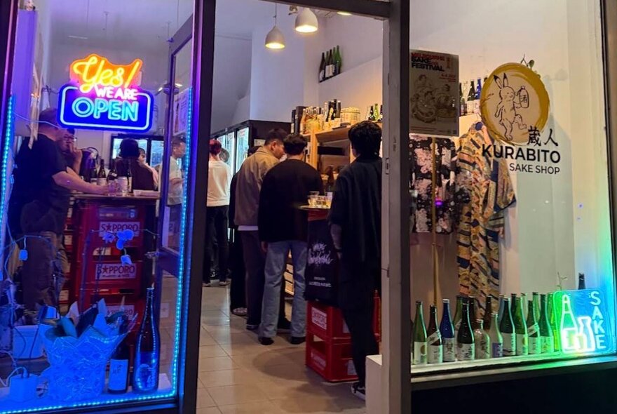 Small Japanese liquor shopfront and bar with neon signs, bottle displays and people seated at the bar and tables.