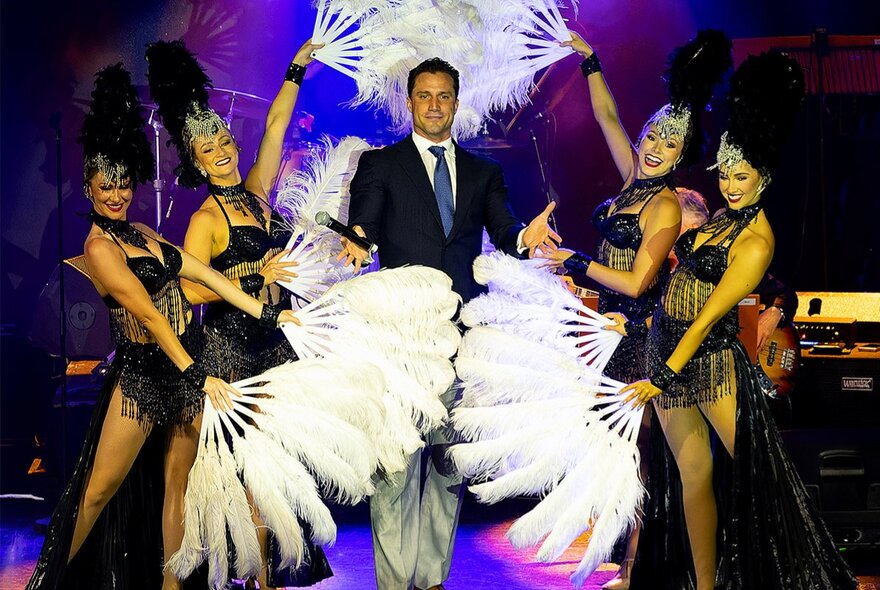 Performer, PJ Lane, on stage in a suit with four burlesque dancers surrounding him with white feather fans.