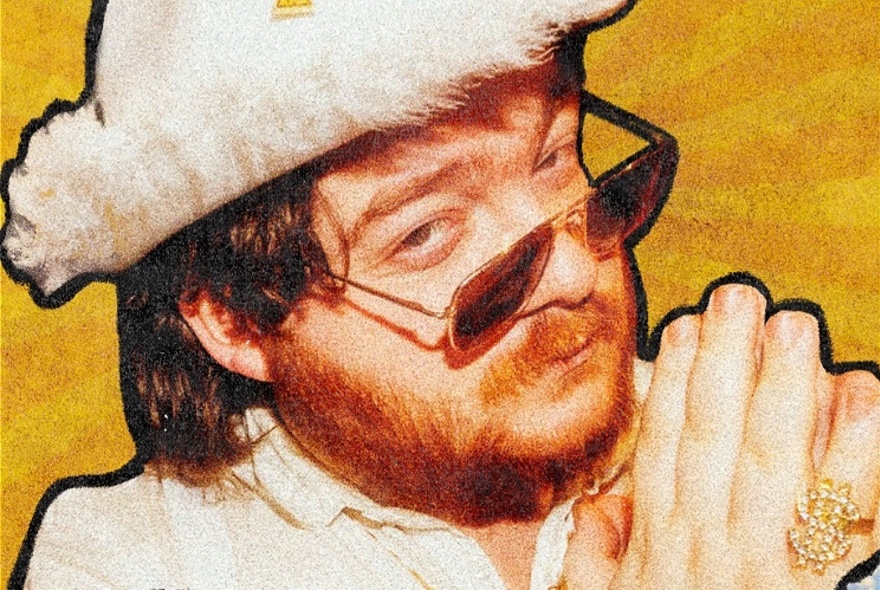 A man in a white feathered cowboy hat, sunglasses and a white suit, with palms together as though praying.