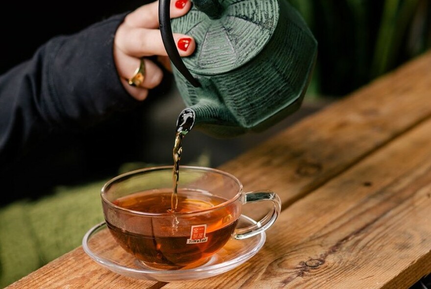 A woman's hand pouring tea from a green teapot into a clear cup and saucer.