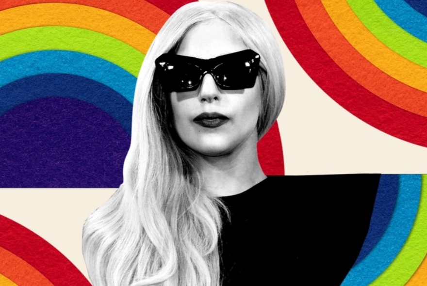 A black and white image of Lady Gaga wearing dark sunglasses, against a rainbow coloured backdrop.