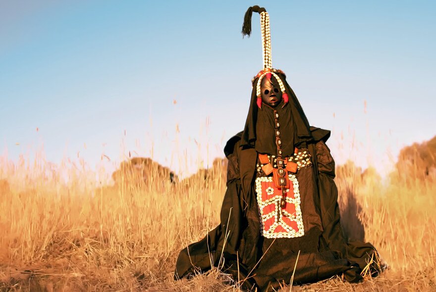 A person in a dry landscape wearing an African tribal-inspired outfit with mast and tassel on top.