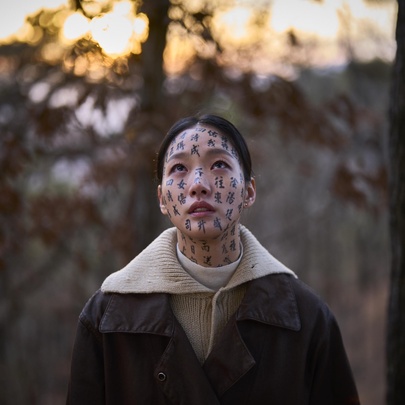 Close-up of actress Kim Go-eun from the film Exhuma, looking upward with protective spiritual characters written across her face in a forest setting at sunset.