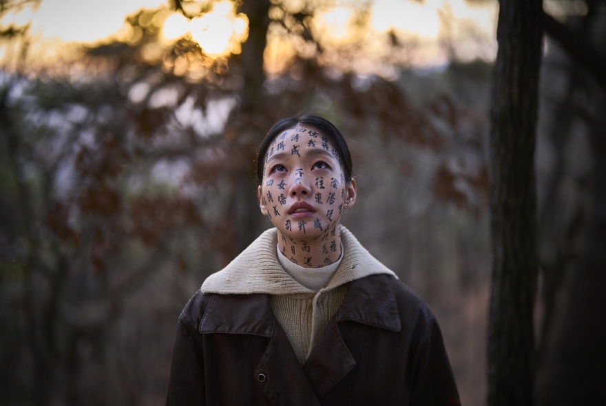 Close-up of actress Kim Go-eun from the film Exhuma, looking upward with protective spiritual characters written across her face in a forest setting at sunset.