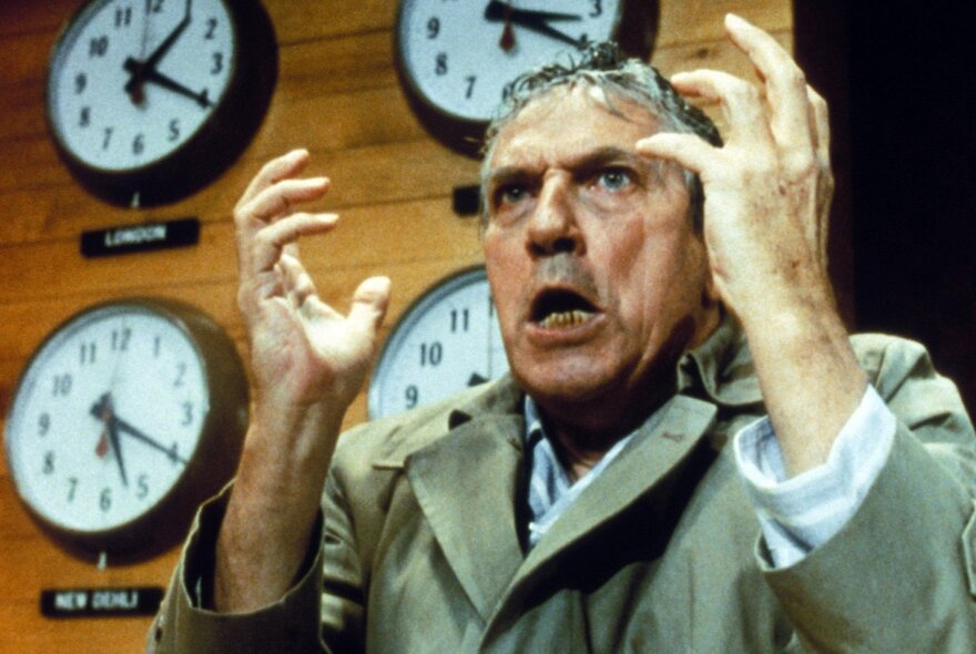 A still from the film, Network, showing a man in a trench coat with an exasperated expression, in front of four clocks mounted on a wooden wall.