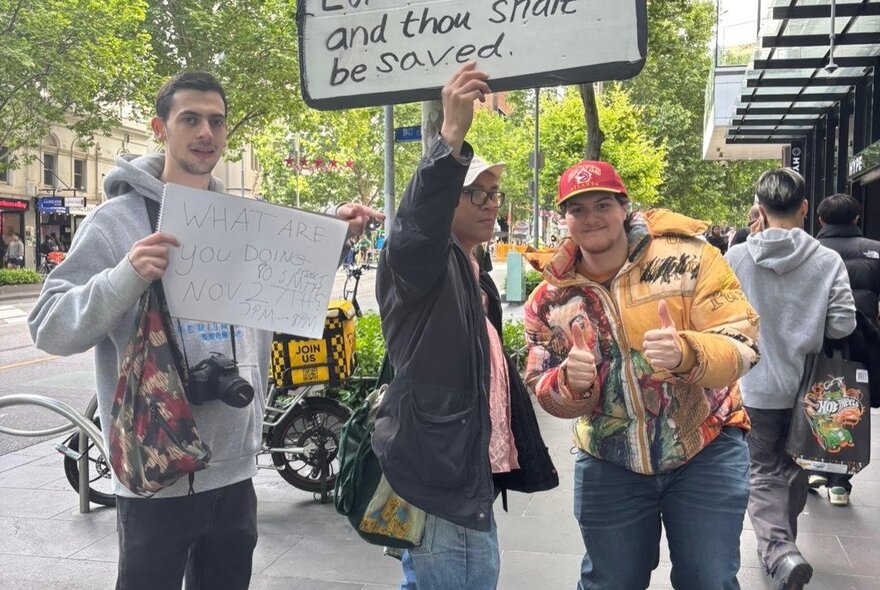 Three people interacting on a Melbourne city street, two of them holding up signs, with trees, a bike and passers-by in the background.