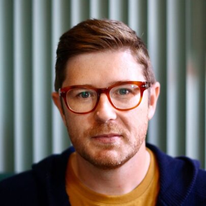 Portrait of Canadian political commentator, David Moscrop, wearing red glasses, a mustard t-shirt and an open blue hooded jumper.