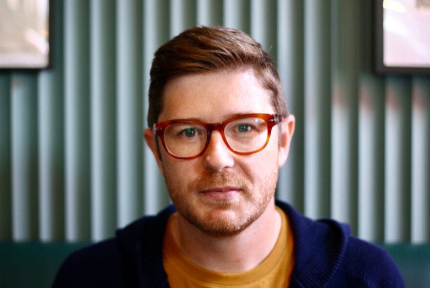 Portrait of Canadian political commentator, David Moscrop, wearing red glasses, a mustard t-shirt and an open blue hooded jumper.