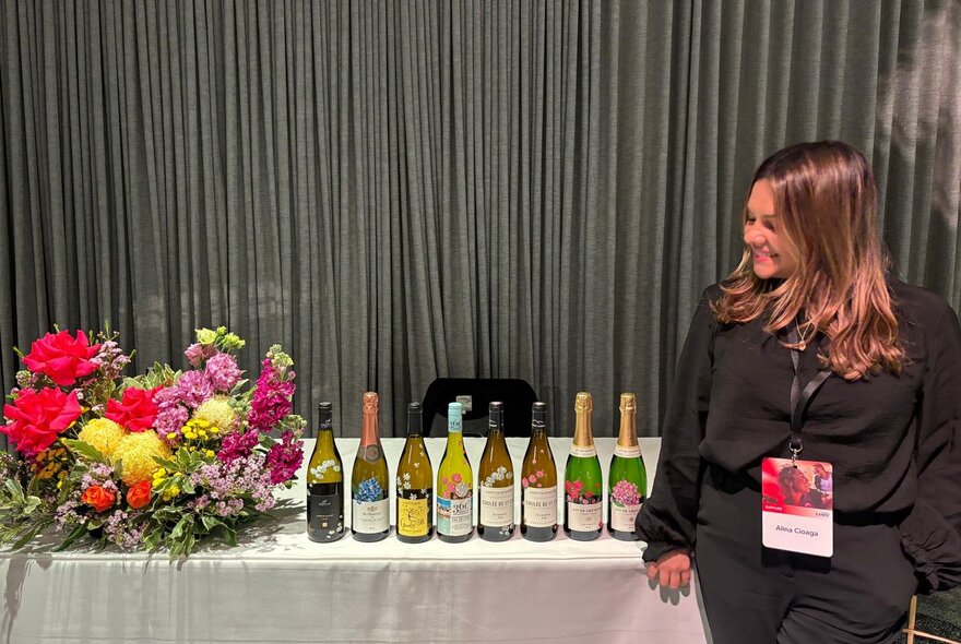 Person standing next to a row of wine bottles and a vase of flowers on a display table.