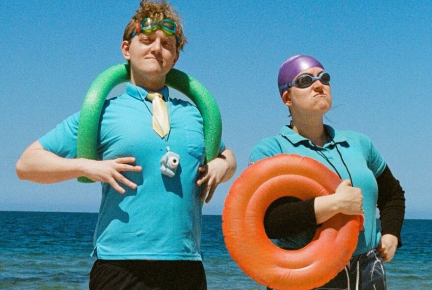 Two actors wearing aqua t-shirts, both with swimming goggles and inflatable pool devices striking a pose with the sea as the backdrop.