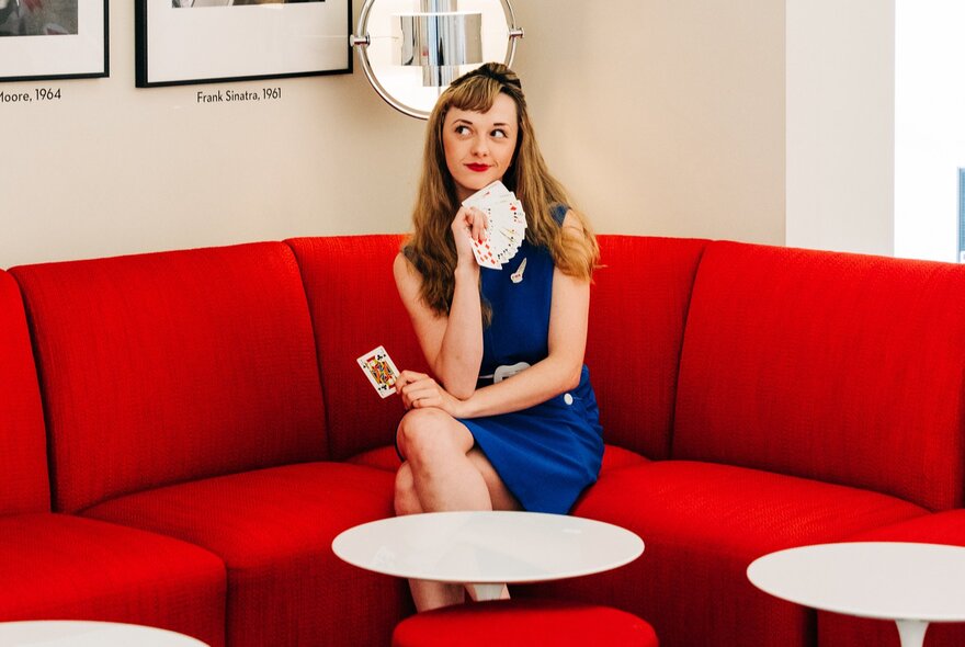 Performer Magical Katrina wearing a blue dress, holding playing cards in her hand, seated on a red couch.