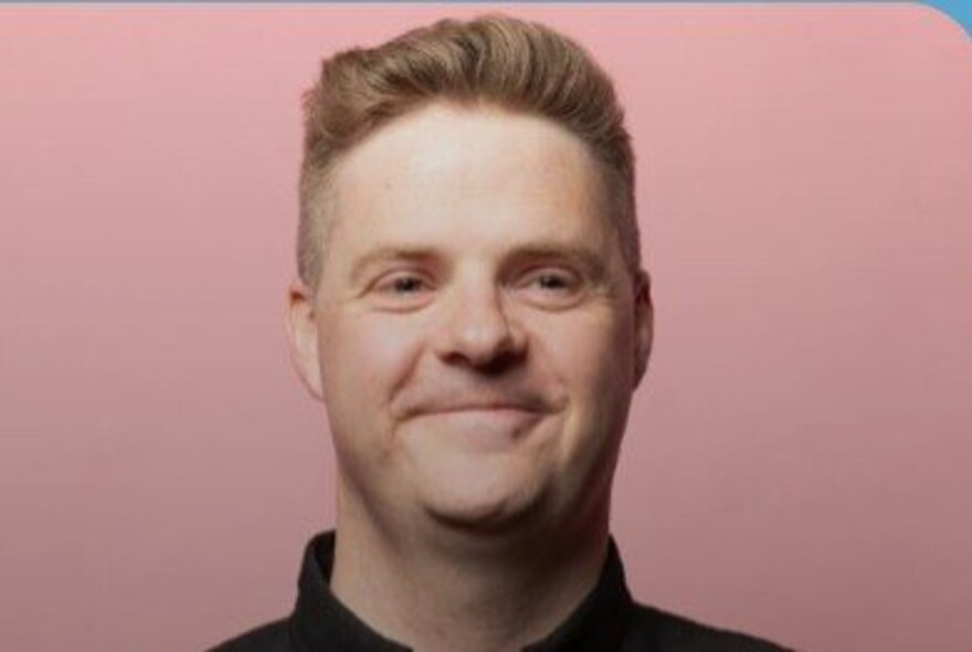 Portrait shot of Australian comedian, Tom Ballard, smiling at the camera; pink background. 