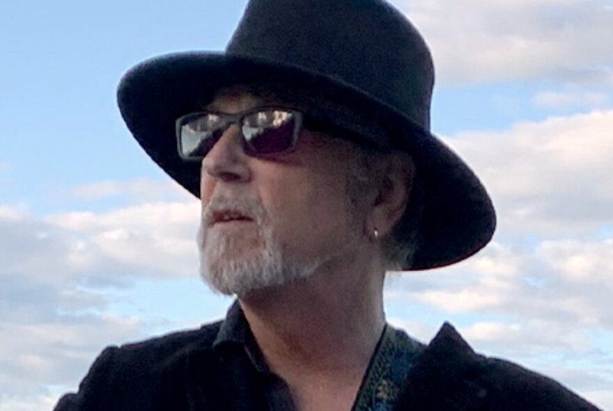 Singer and musician Steve Kilbey wearing a large black hat and sunglasses, looking wistfully at the sky. 