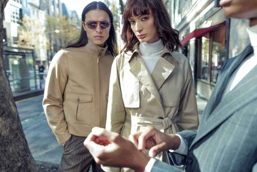 Three models, one wearing a bomber jacket, one in an overcoat and one wearing a suit jacket,  standing in a city street outside shopfronts that are just visible behind them.