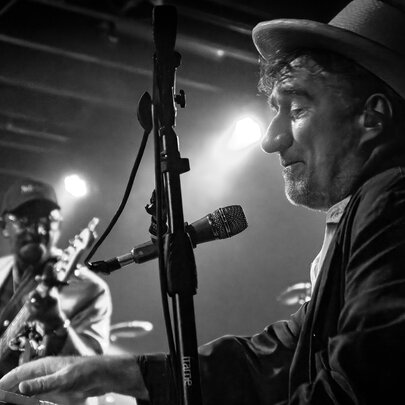 Musician Jon Cleary wearing a hat playing keyboards live on stage, with a guitarist in the background; black and white image.