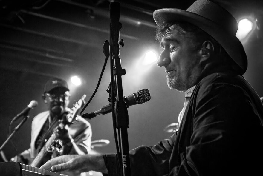 Musician Jon Cleary wearing a hat playing keyboards live on stage, with a guitarist in the background; black and white image.