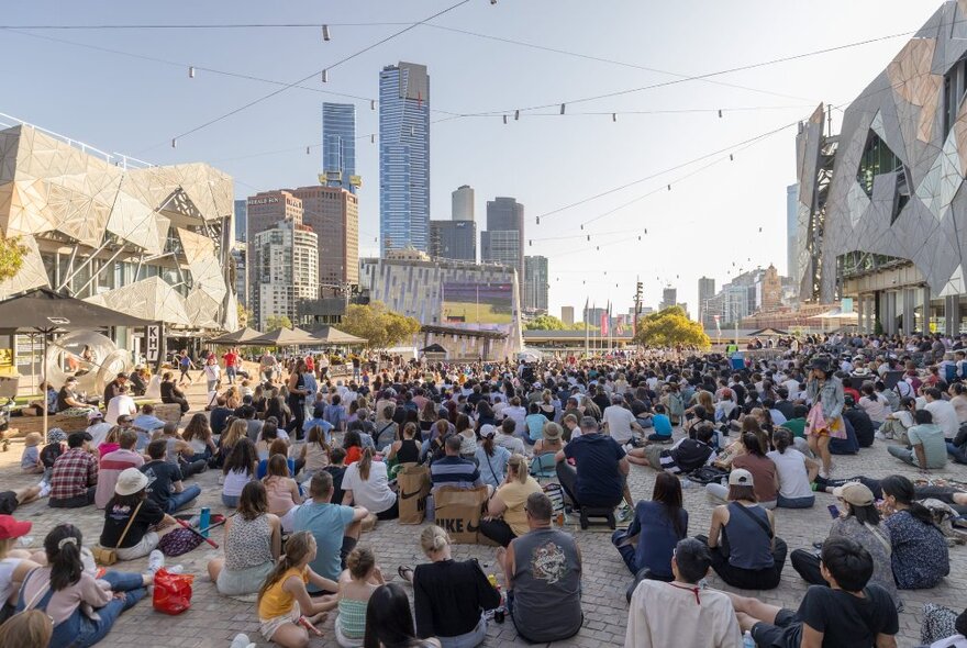 A crowd sitting watching the Big Screen and Federation Square in Melbourne at dusk. 