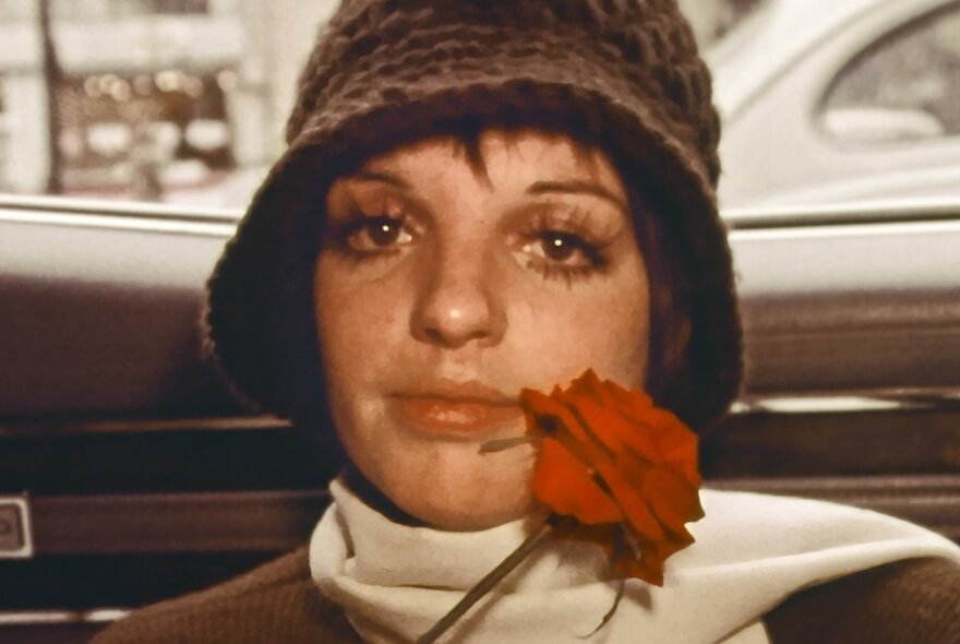 Film still of the actress Liza Minelli wearing a hat nd scarf and holding a red rose.