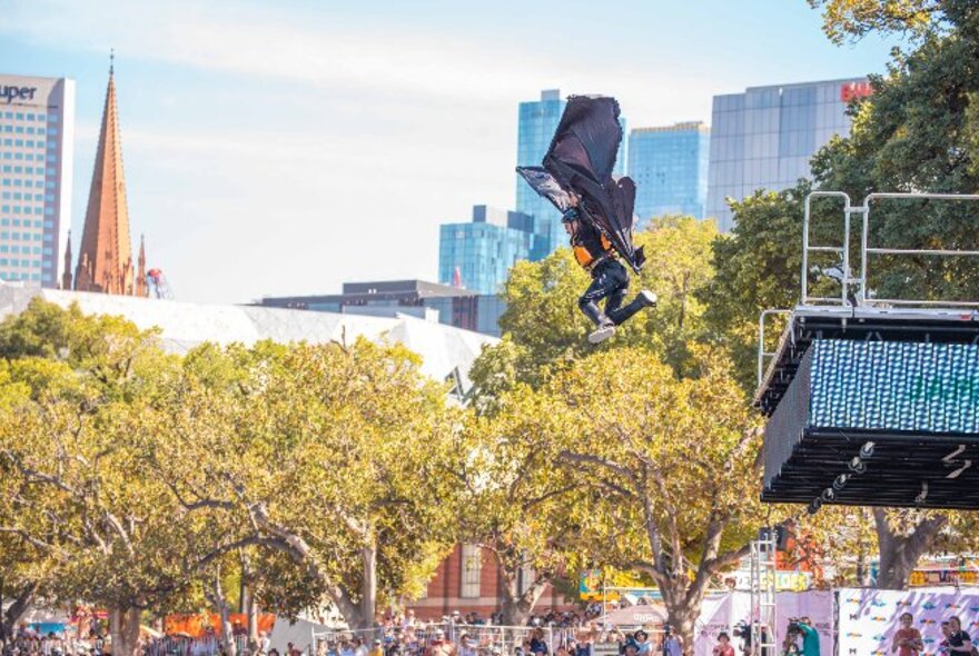 A person in a home made flying suit leaping off a platform, with Melbourne city skyline and trees in the background.