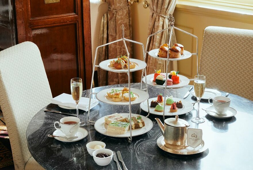 Afternoon tea with little plates of savoury and sweet food stacked up above each other on two three-tiered stands, on a table with pots of tea, cups and glasses of sparkling wine.