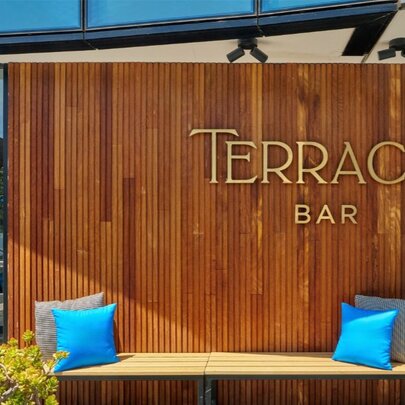 A wood-panelled exterior wall featuring a brass sign that reads "Terrace Bar". Below the sign is a light-coloured bench decorated with two bright blue throw pillows.