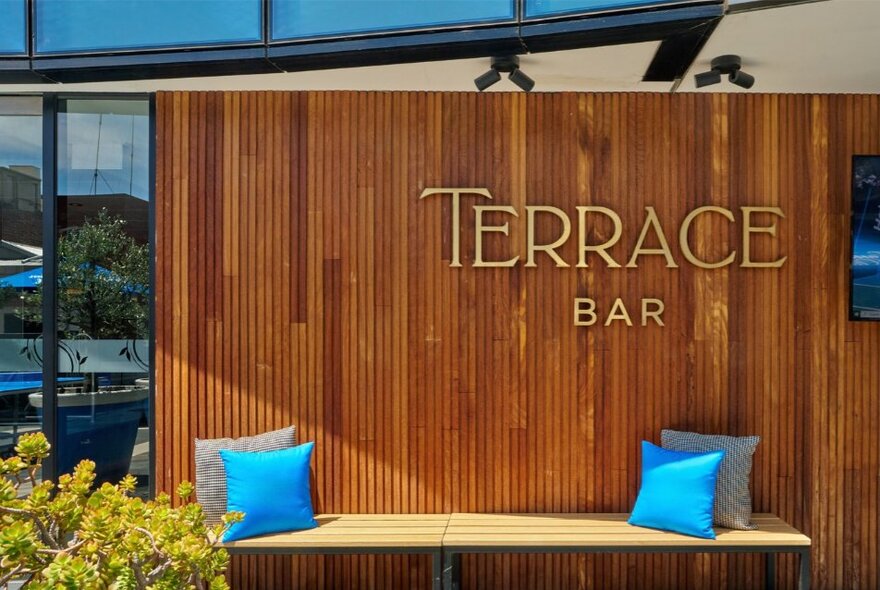A wood-panelled exterior wall featuring a brass sign that reads "Terrace Bar". Below the sign is a light-coloured bench decorated with two bright blue throw pillows.