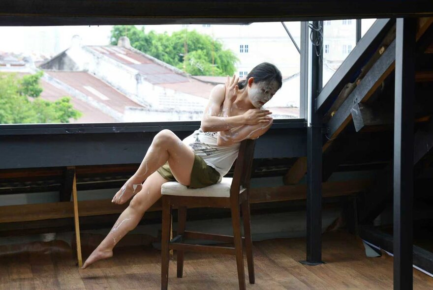 A solo dance performance on a balcony with a woman on a chair with white face makeup. 