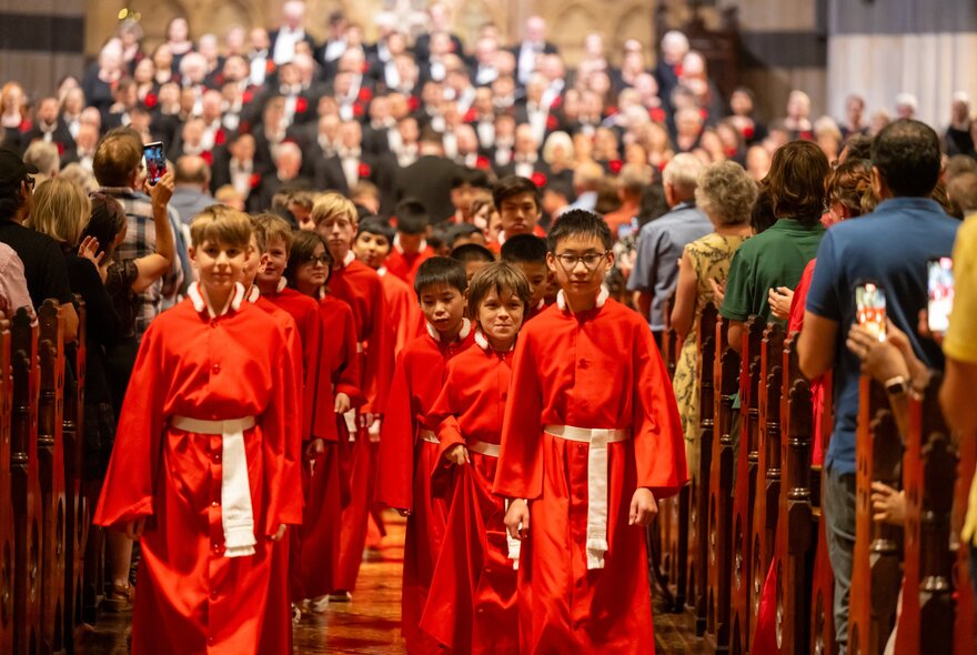 Choir boys dressed in red walking down the central aisle of a cathedral, with a standing choir in the background that are out of focus, and people seated in pews on either side of the aisle.