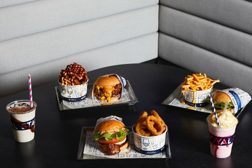 Trays of food on a table containing hamburgers, onion rings, french fries and shakes.