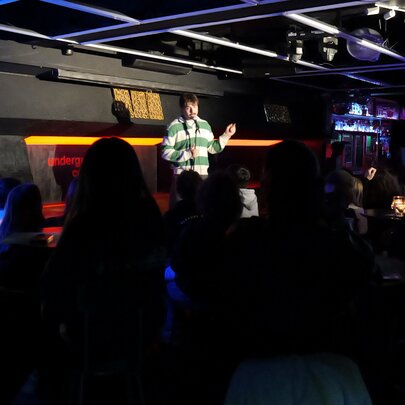 Stand-up comedian standing on a stage holding a microphone in front of a small seated audience.