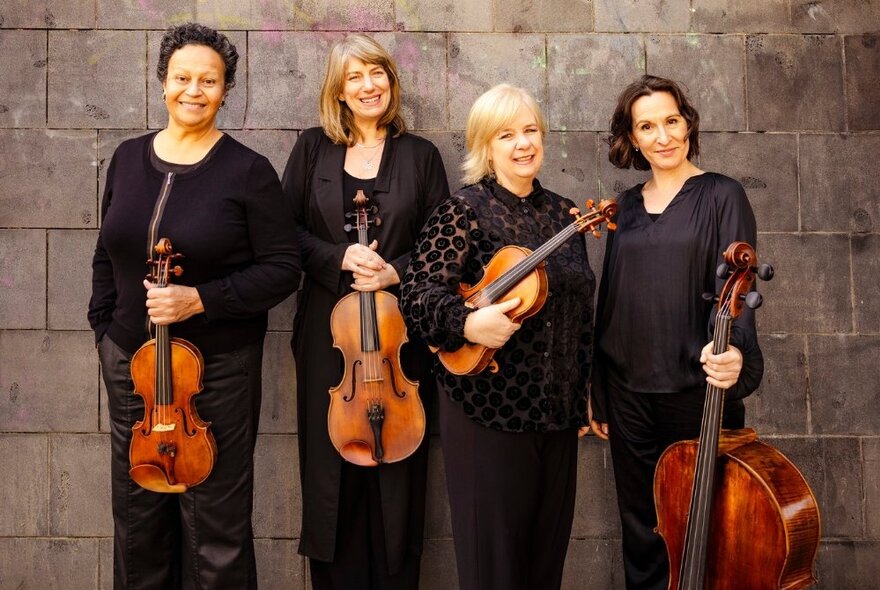 Four female classical musicians holding stringed instruments, wearing black, standing against a grey brick wall.