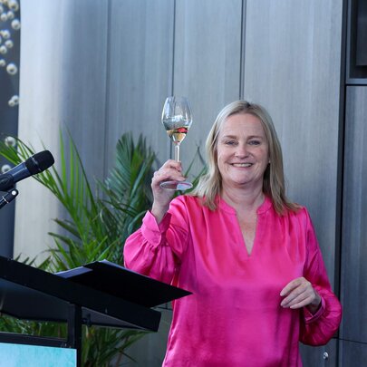 A woman in a bright pink dress raised a glass of champagne and smiles openly in a warehouse type setting with a microphone close by.