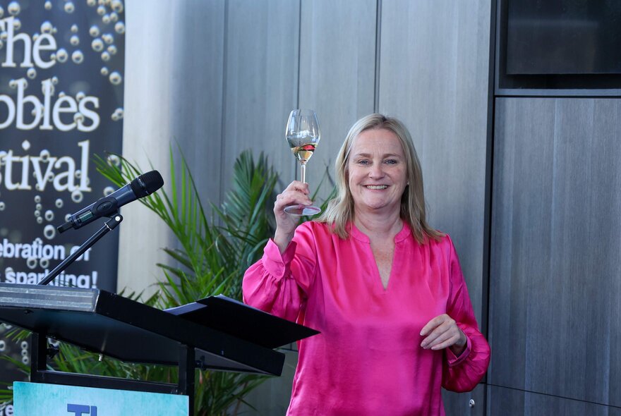 A woman in a bright pink dress raised a glass of champagne and smiles openly in a warehouse type setting with a microphone close by.