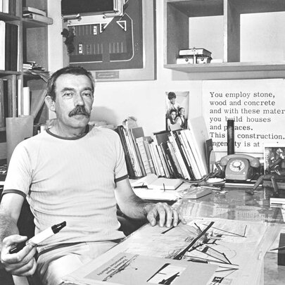 Black and white image of architect Enrico Taglietti, sitting at his studio desk, surrounded by books and papers.