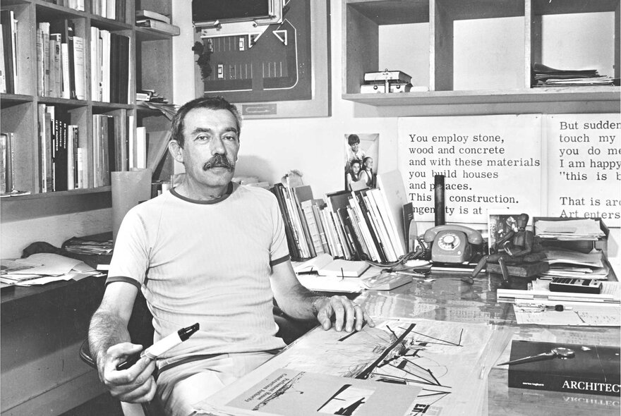 Black and white image of architect Enrico Taglietti, sitting at his studio desk, surrounded by books and papers.